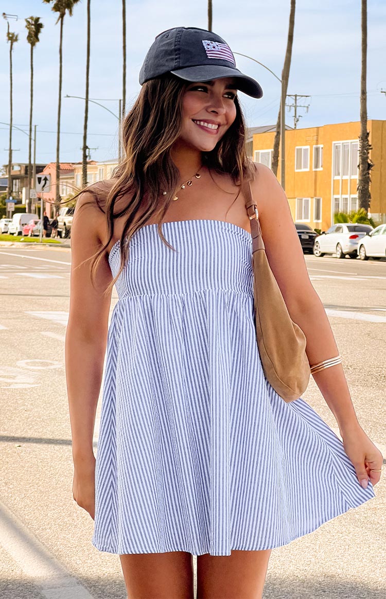 Model is wearing a blue and white pinstripe strapless mini dress with a babydoll silhouette. She has paired it with a blue baseball cap and a tan suede shoulder bag.