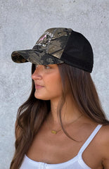 Model wearing a green and brown camo printed trucker hat with a pink pony embroidered graphic