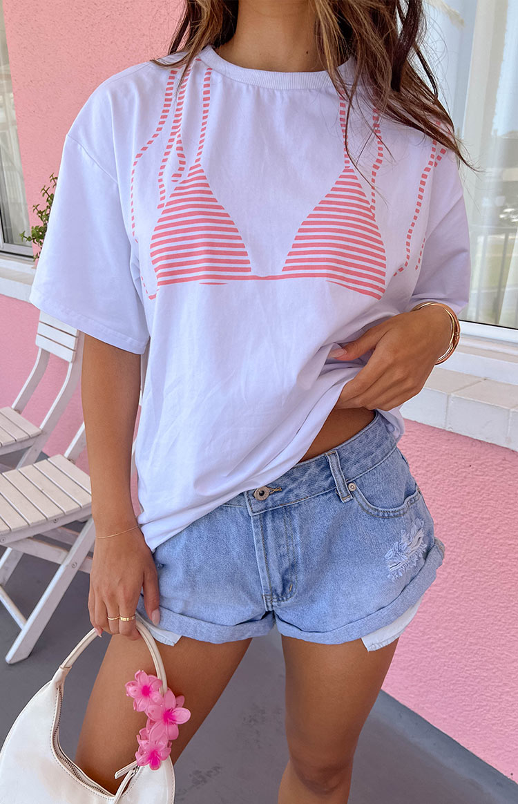 Girl wearing a white oversized tee with a pink bikini top graphic printed on it. Wearing denim cuffed shorts and holding a white handbag with a pink flower hairclip.