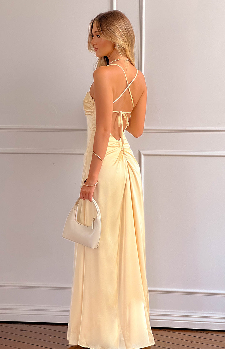 blonde model wearing a yellow satin maxi dress with an open back and strap details holding a white handbag