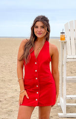 Model is wearing a vibrant red denim mini dress designed with a halter neck and prominent buttons down the front. The dress also features two small flap pockets.