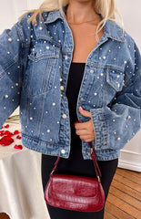 a girl with blonde hair wears a denim jacket with peals all over, a black dress and gold necklace and holds a red purse