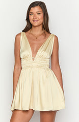 Brunette model wearing a yellow satin mini dress and gold jewellery holding a beige woven handbag.