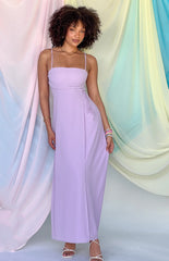 Brunette girl in lilac maxi dress standing in front of pastel curtains.