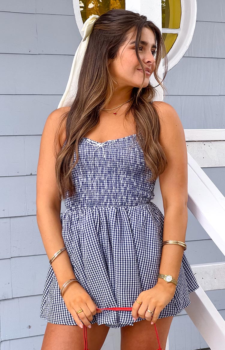 Model is wearing a navy and white gingham playsuit featuring a shirred bodice- thin shoulder straps- and a small white bow detail at the neckline. Her hair is accented with a cream bow.