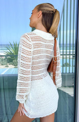 Girl facing away wearing white crochet mini dress and brown shoulder bag in front of glass door.