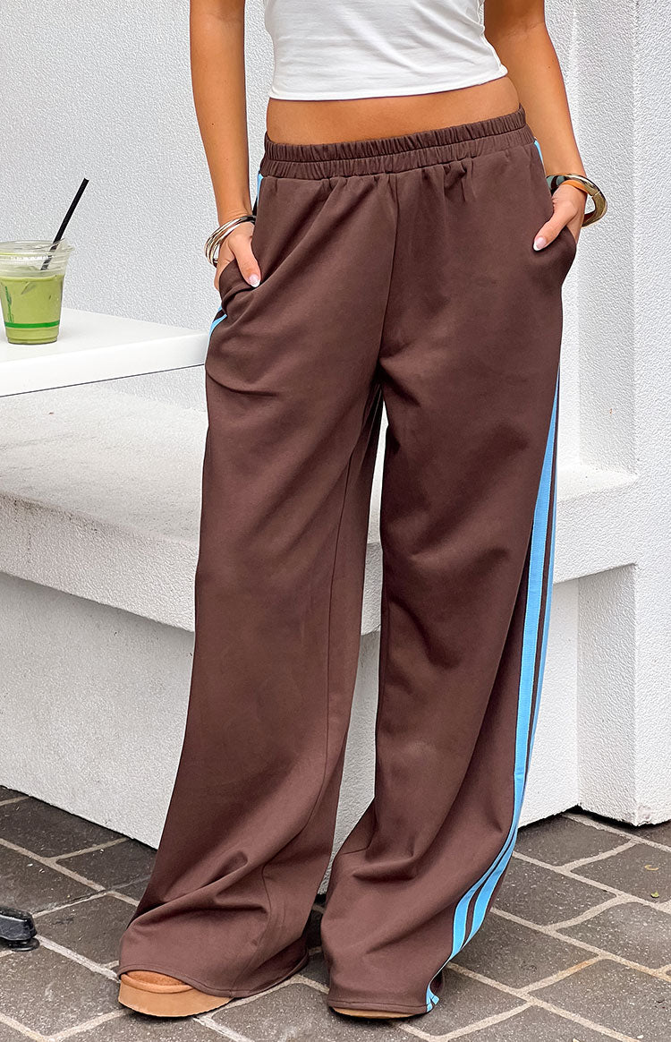 Full-length image of a person wearing a pair of brown wide-leg track pants with a light blue stripe down each side. Our pants have an elastic waistband and pockets best for your athletic mood.