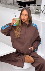 A female model wearing an oversized brown zip-up track jacket with a high collar and light blue stripes down the arms.