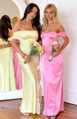 Blonde model wearing a pink satin formal gown with off shoulder straps paired with black heels and a small flower bouquet
