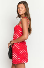 Model wearing a vibrant red mini dress with white polka dots- a sweetheart neckline- and thin tie-straps- accessorized with gold jewelry and a dark bag.
