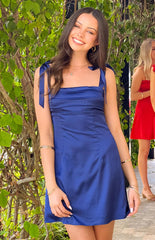 Model smiling in a navy blue satin mini dress- perfect for homecoming. The dress features a straight- pleated neckline and wide shoulder straps that tie into bows.