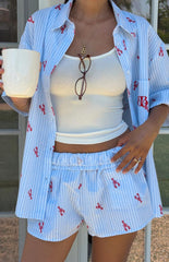 A woman in a light blue and white striped pajama shirt with a red lobster print- unbuttoned over a white ribbed tank top- and matching blue and white striped shorts with the same red lobster print.