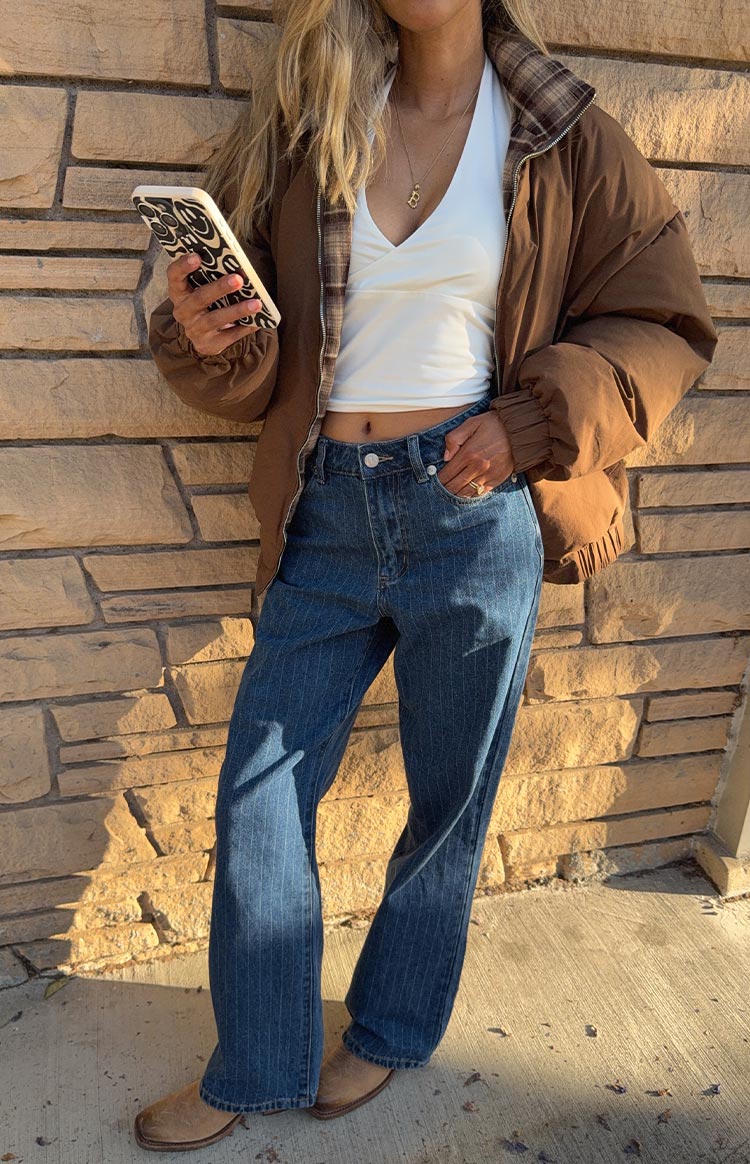 A woman in a white halter neck crop top and dark wash pinstripe wide-leg jeans- with a brown puffer jacket featuring a brown plaid lining