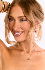 A model wears a delicate gold hoop earring with a red heart pendant. This is a perfect accessory for a date night outfit or a casual day look. The earring features a small polished heart charm.