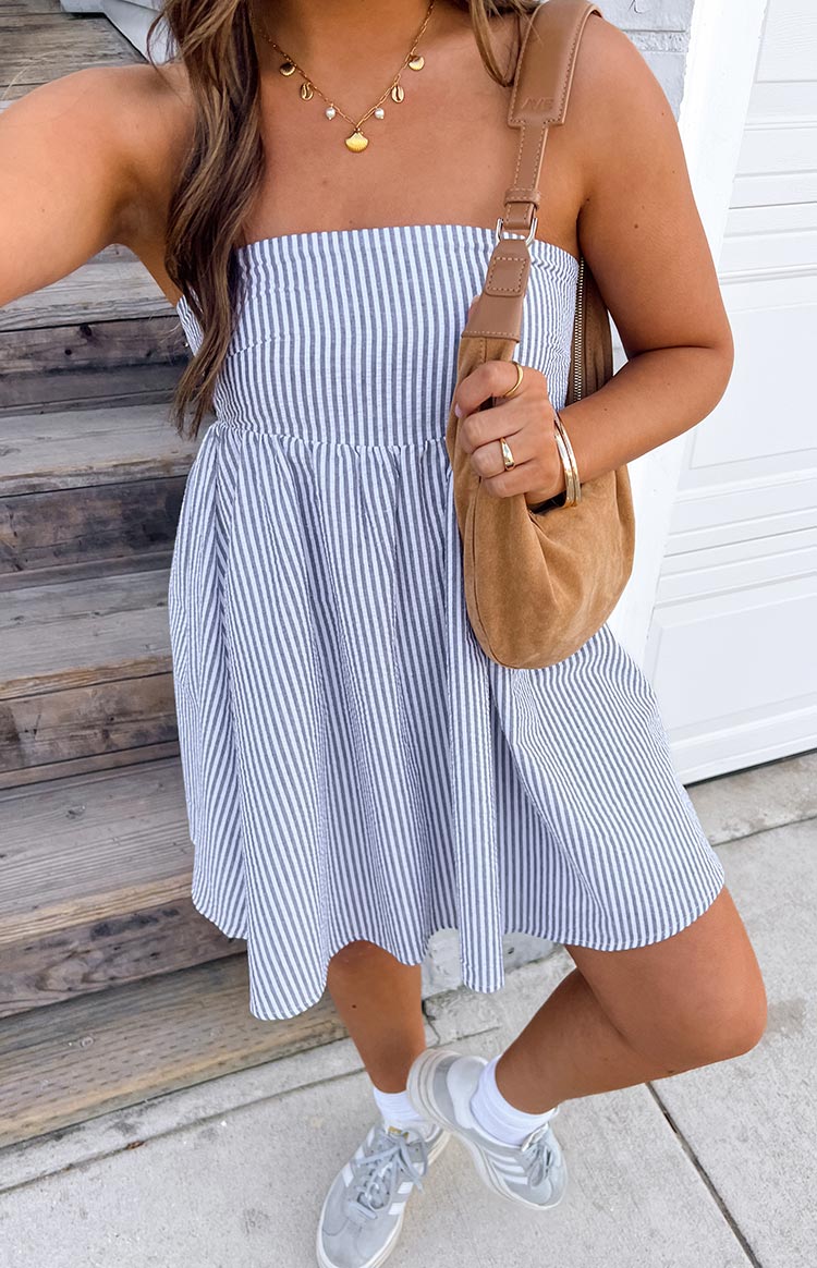 Girl is styled in a blue and white vertically striped strapless mini dress with an empire waist. The dress is accessorised with layered gold necklaces- a tan shoulder bag- and grey sneakers with white socks.