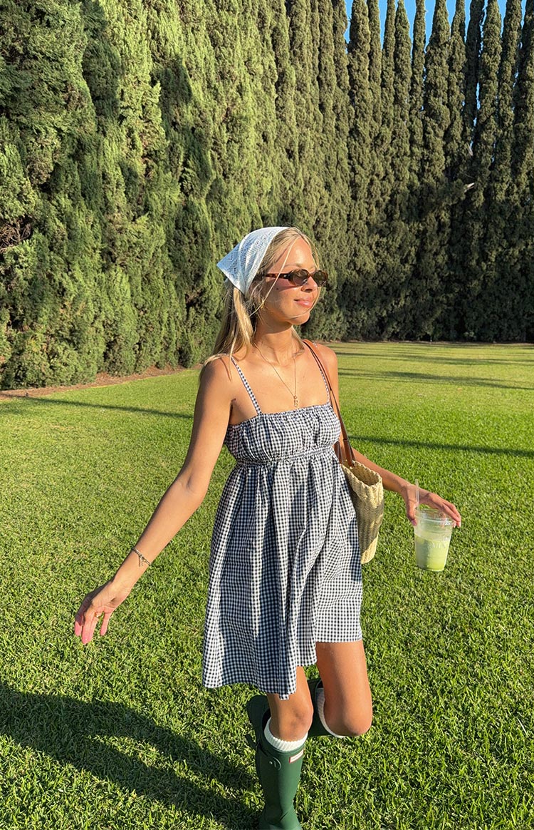 A woman in a black and white gingham mini dress with a ruched bust and spaghetti straps- a white bandana tied around her head- and dark green rain boots with white socks.