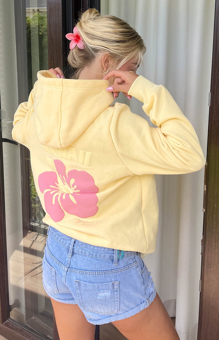 Blonde model is wearing a yellow fleece hoodie with a pink hibiscus flower print on the back. She is wearing blue denim shorts and a pink flower hair clip.