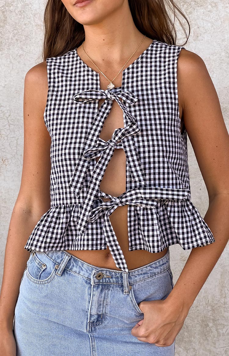 A close-up of a black and white gingham print sleeveless top featuring a deep V-neckline with multiple tie-front closures and a ruffled hem.