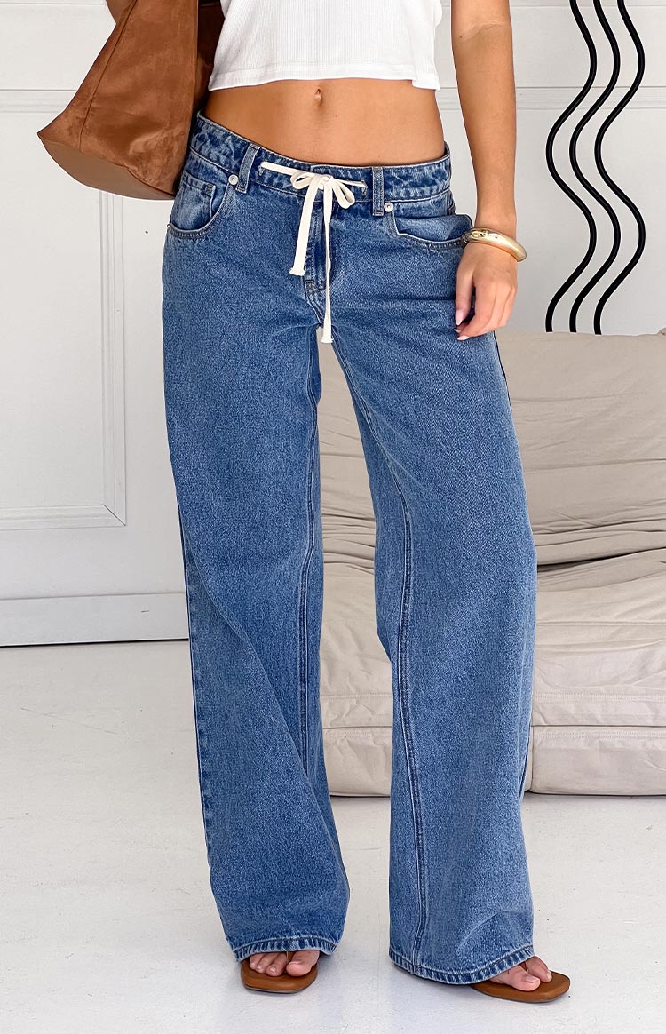 Model wearing stylish wide-leg blue denim jeans with a drawstring waist- paired with a white crop top and brown accessories.