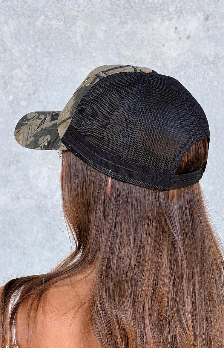 Model wearing a green and brown camo printed trucker hat with a pink pony embroidered graphic