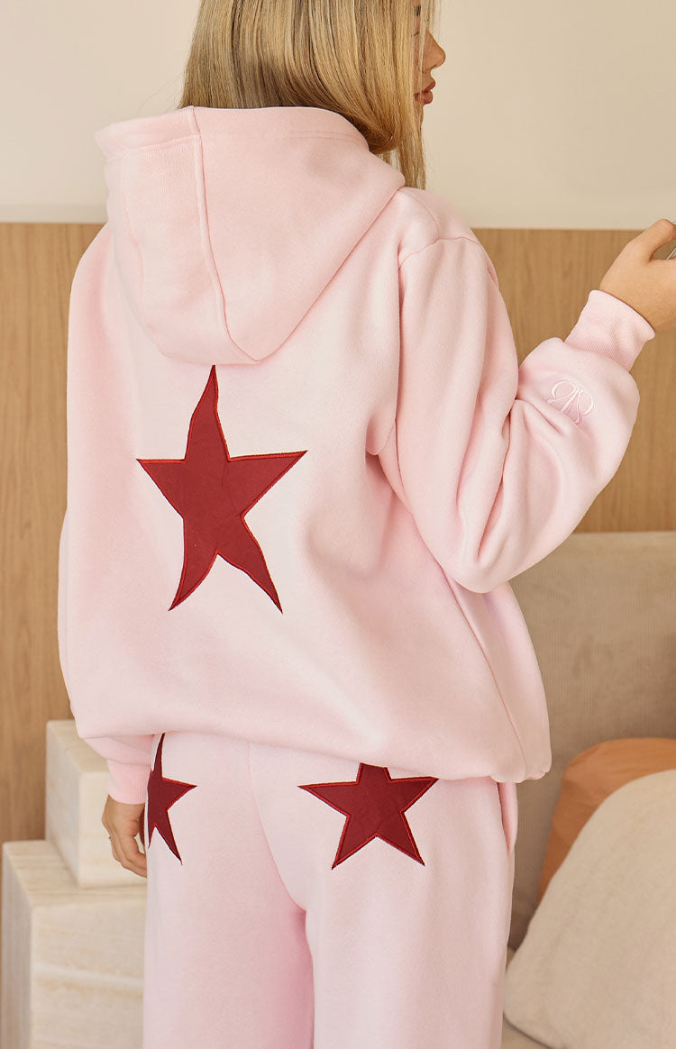 The back view of a light pink hoodie featuring a large dark red star applique and a small embroidered detail on the sleeve cuff.
