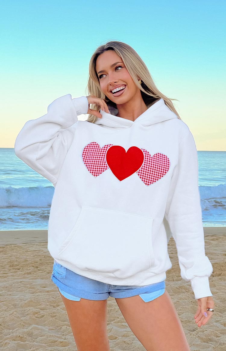 A white hooded sweatshirt featuring three heart appliqués on the front: a solid red heart in the center flanked by two red and white gingham hearts.