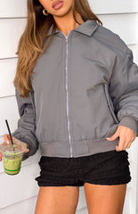 A female model wearing a grey bomber jacket with a ribbed collar cuffed sleeves and a zip-up front.