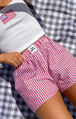 Model is wearing red and white vertically striped boxer-style shorts with an elasticated waistband. The shorts are paired with a white ribbed cropped tank top that has an American flag embroidery.