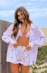 Model is wearing a white oversized long-sleeve shirt with a scattered red strawberry embroidery pattern- open to reveal a red and white gingham bikini top- paired with matching white strawberry embroidered boxer shorts.