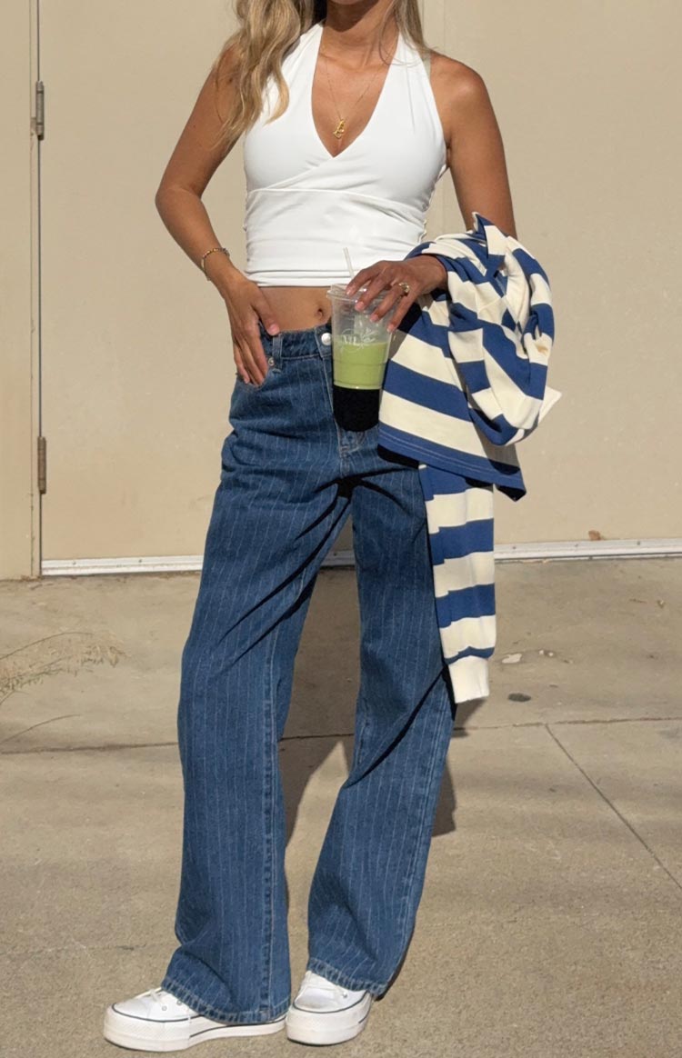 A woman wears a white halter neck crop top and mid-wash- mid-rise slouchy jeans with subtle pinstripes. She holds a blue and white striped long-sleeve rugby top over her arm and wears white canvas platform sneakers.