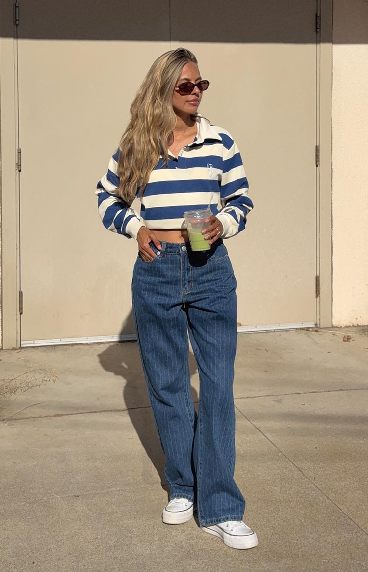 A woman wears a white and blue striped long-sleeve cropped rugby top with a white collar- paired with mid-wash- mid-rise slouchy jeans with subtle pinstripes- and white canvas platform sneakers.