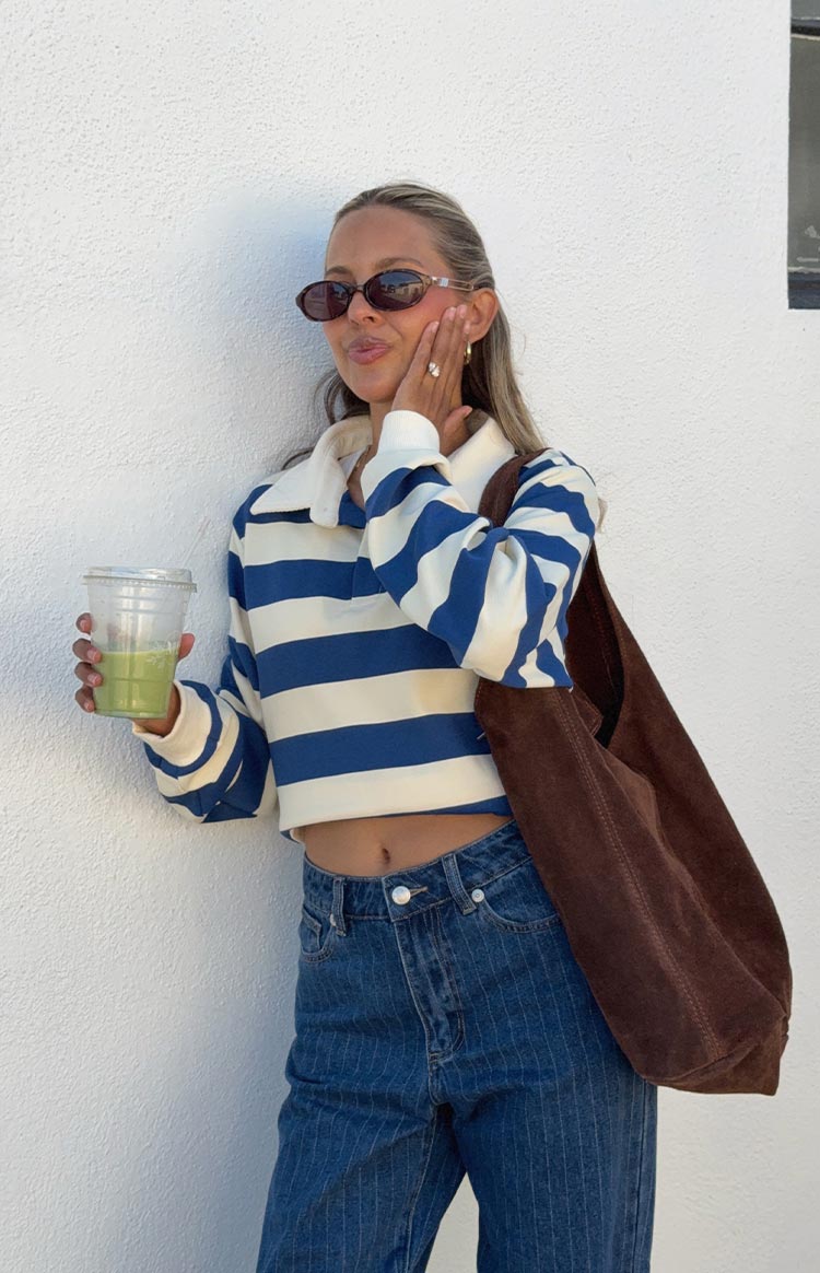 A woman in a cropped- long-sleeve rugby top with wide cream and blue horizontal stripes and a white collar- paired with mid-wash- mid-rise slouchy jeans with subtle pinstripes.
