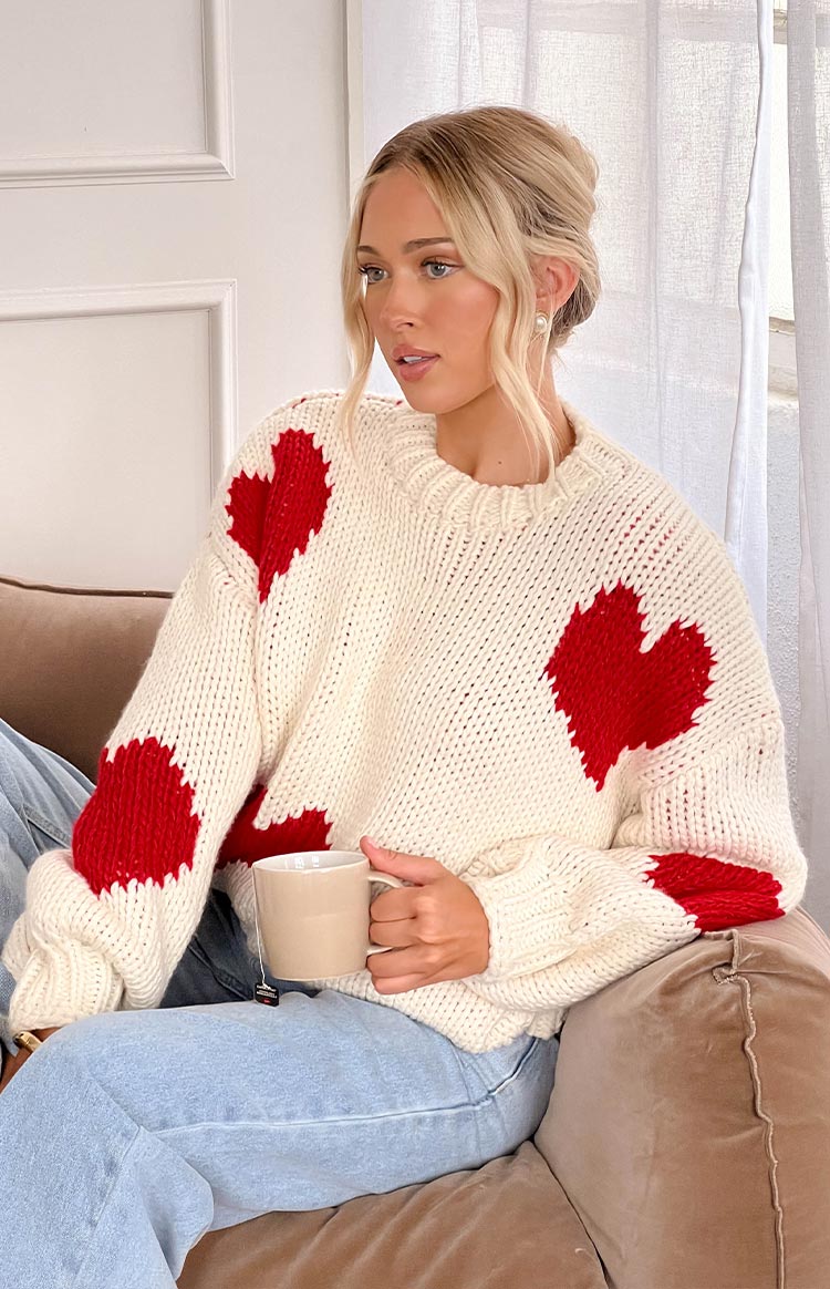 a white chunky knit sweater with red hearts is worn by a blonde girl with denim jeans, holding a mug sitting on the couch