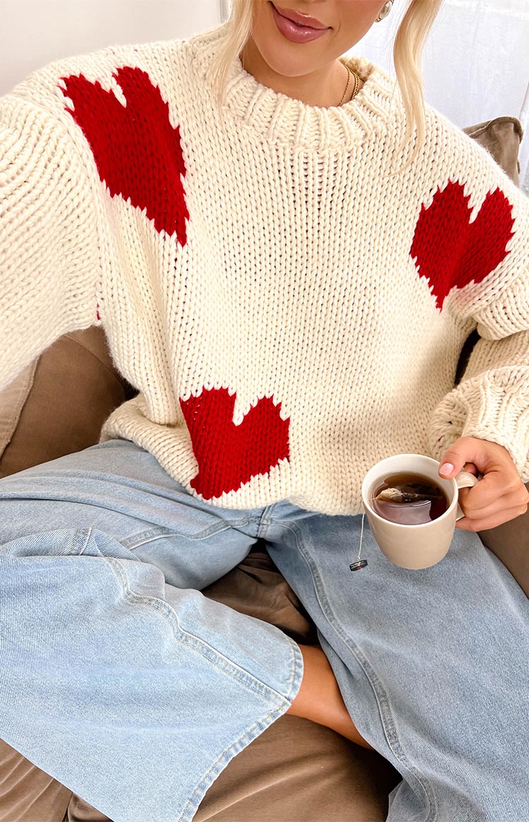 a blonde girl wears a white knit sweater with red hearts, denim jeans and holding a cup of tea