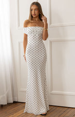 A female model wearing a white off-the-shoulder maxi dress with a black polka dot pattern. The dress features a ruched bodice and a straight floor-length skirt.
