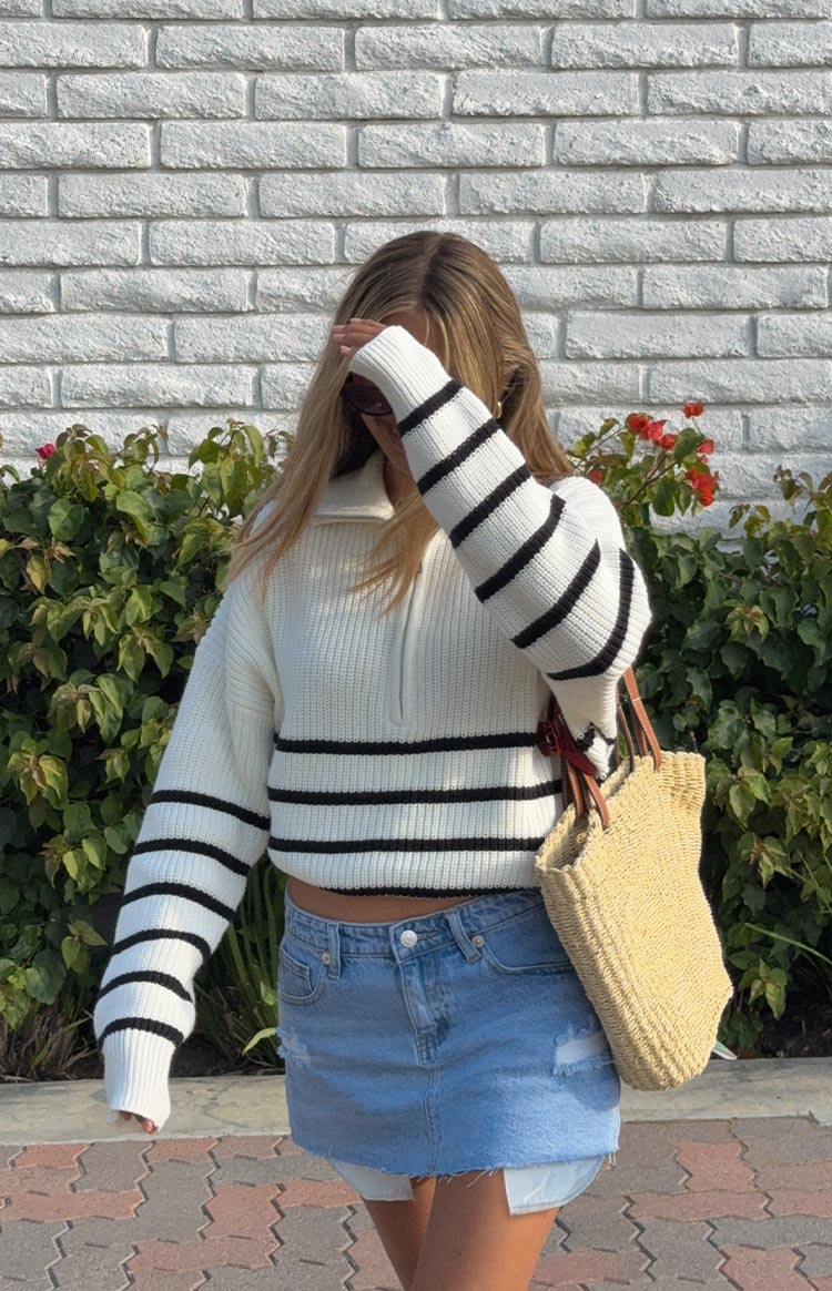 A woman wears a cream knit quarter-zip jumper with black horizontal stripes on the body and sleeves- and a light wash denim mini skirt