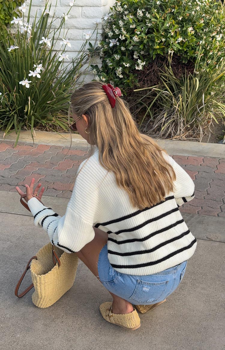A woman from behind- wearing a cream knit quarter-zip jumper with black horizontal stripes on the body and sleeves- paired with light wash denim cutoff shorts and straw-colored flat shoes with a gold buckle.