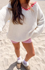 Girl is styled in an oversized cream chunky knit sweater featuring red contrast stitching along the V-neck and cuffs. She wears it with red and white striped shorts and beige shearling-lined slippers over white socks with red heart details.
