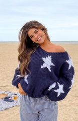 Model is wearing a navy blue chunky knit sweater adorned with large white knitted star patterns on the body and sleeves. The sweater has a wide neckline and is paired with navy and white horizontally striped pants.