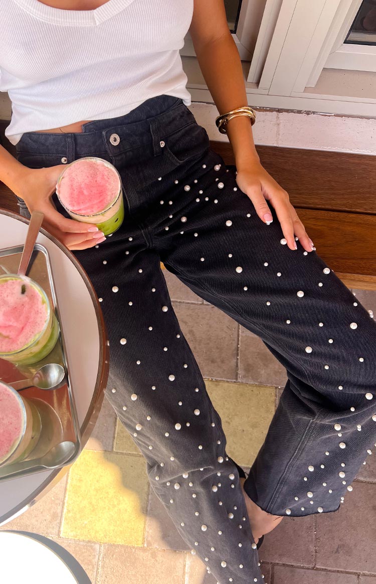 a blonde model sits wearing a pair of black denim jeans with pearl detailing and a white top holding a strawberry matcha