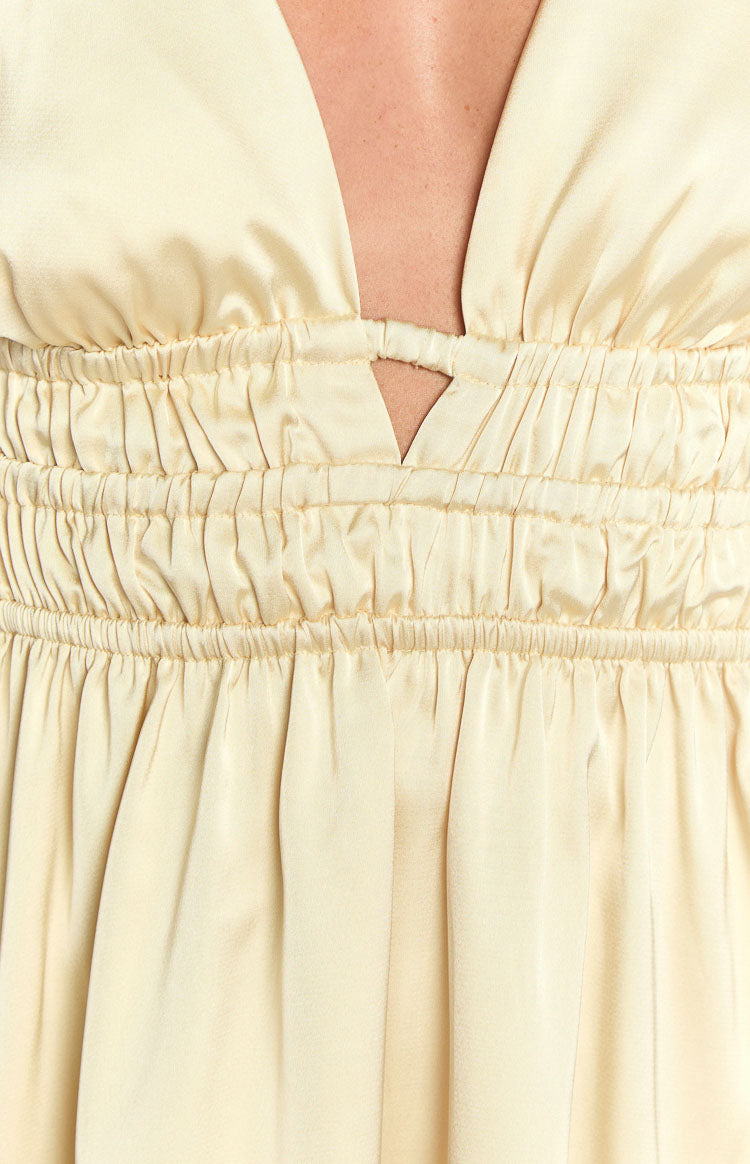 Brunette model wearing a yellow satin mini dress and gold jewellery holding a beige woven handbag.