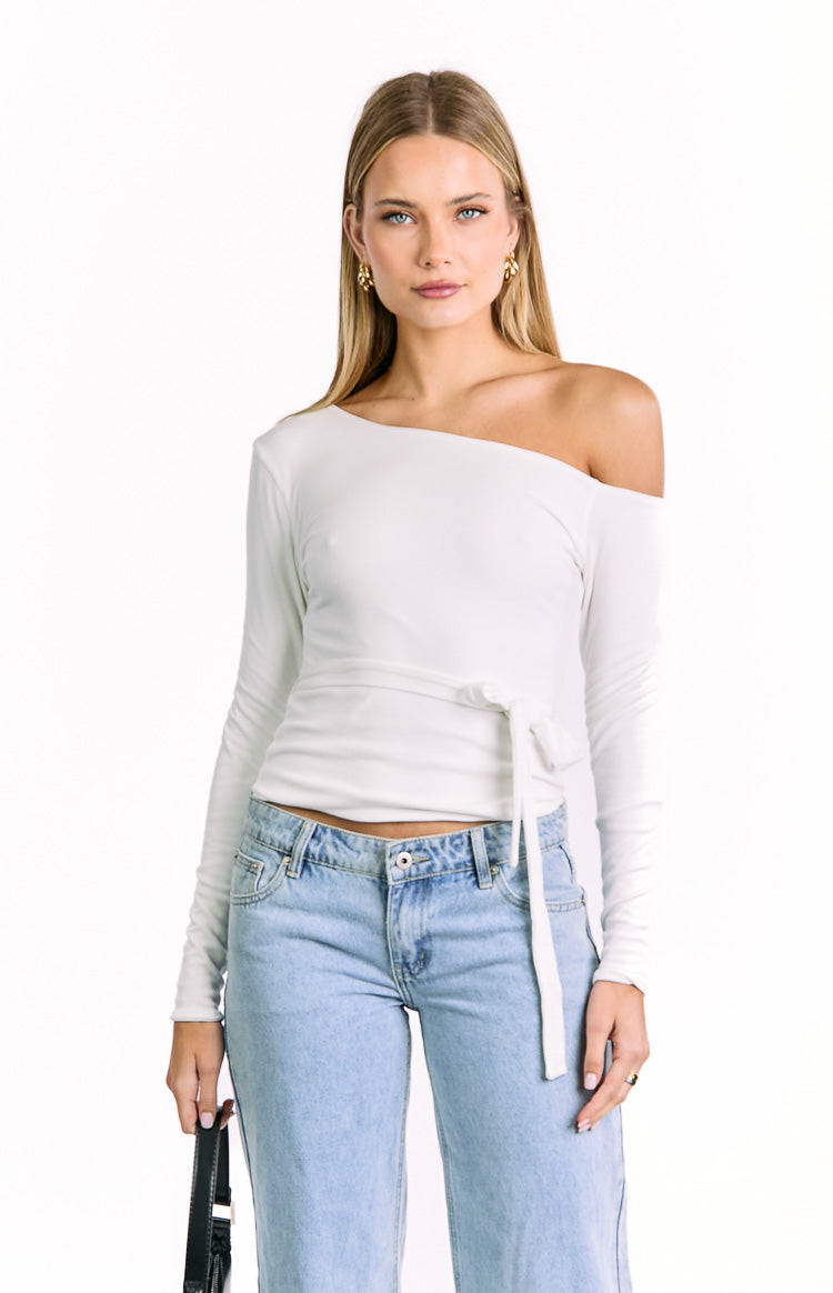 ribbed off-white top with a single-shoulder neckline and a tie closure that wraps around the waist. It is paired with light-colored jeans.
