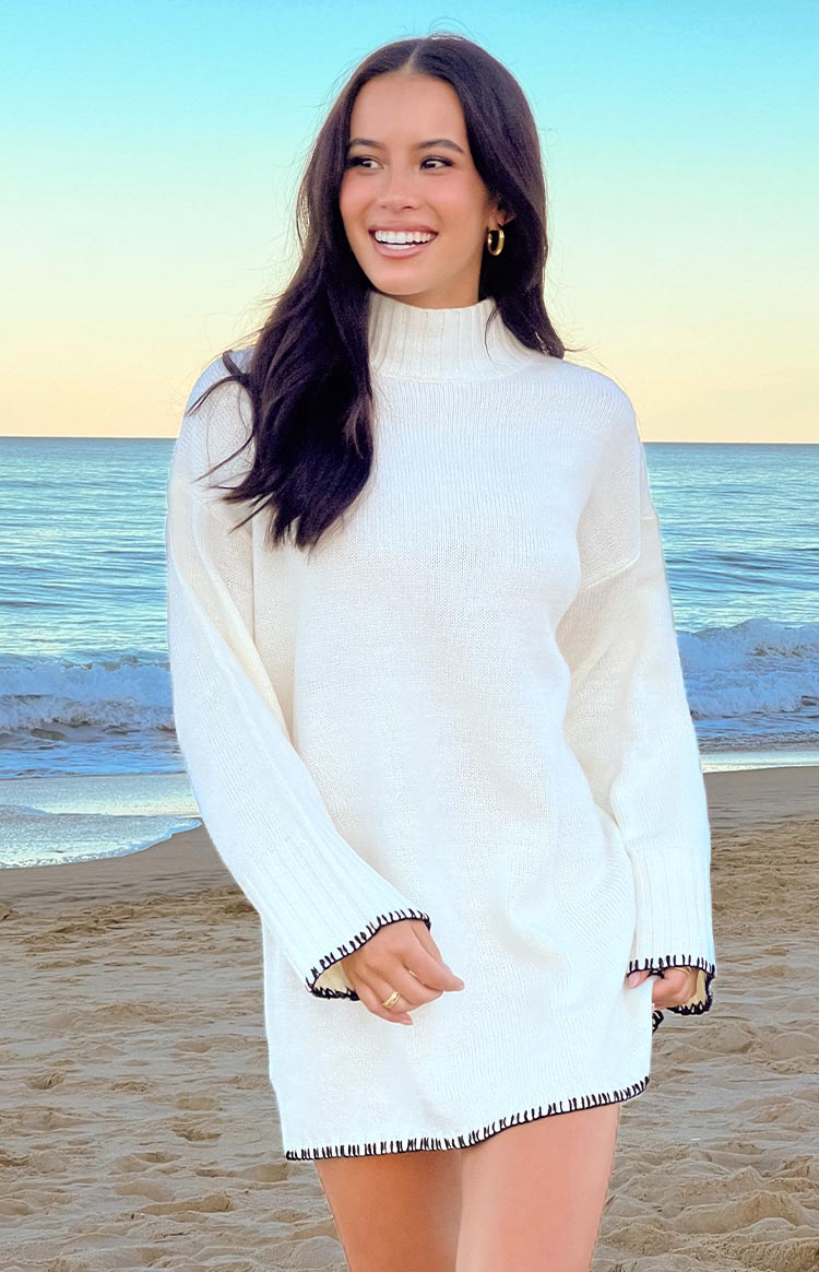 A long-sleeved- relaxed-fit off-white sweater dress with a mock neck and subtle dark contrasting stitching along the hem and cuffs- seen in a full-body shot.