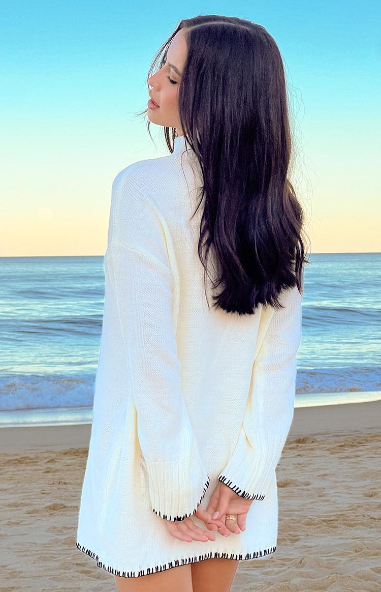 A long-sleeved- relaxed-fit off-white sweater dress with a mock neck and subtle dark contrasting stitching along the hem and cuffs- seen in a full-body shot.