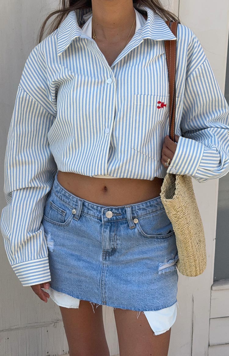 A close-up shot of a person's outfit- featuring a cropped- long-sleeve button-up shirt in a light blue and white vertical stripe pattern with a small red embroidered detail on the pocket. They are wearing a light-wash- low-rise denim mini skirt with distressed details and a frayed hem.