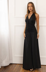 A female model wearing a black halter maxi dress with a plunging V-neckline and a ruched bodice. The skirt is loose and flowing and the model is also wearing black heeled sandals.