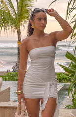 Model is wearing a white strapless mini dress, and a gold seashell earrings. She is holding a beige bag and wearing sunnies.