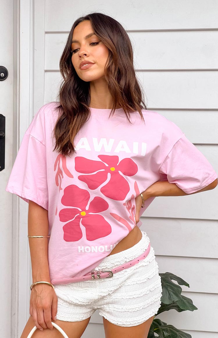 Model with brunette hair wearing a pink oversized shirt and white shorts. She is standing in front of a house.