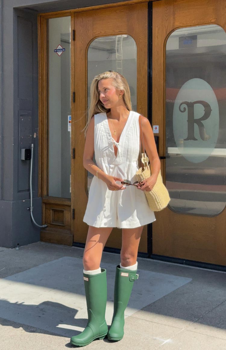 A woman stands outdoors in a sleeveless white playsuit featuring a V-neck with a tie at the bust and vertical pintuck detailing. She is wearing tall- matte green rain boots over white socks that peek out at the top.