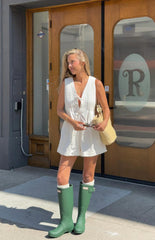 A woman stands outdoors in a sleeveless white playsuit featuring a V-neck with a tie at the bust and vertical pintuck detailing. She is wearing tall- matte green rain boots over white socks that peek out at the top.
